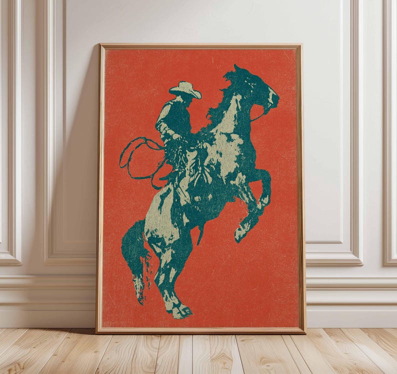 May include: A framed print featuring a cowboy riding a bucking bronco. The artwork uses a teal and off-white color palette against a vibrant orange background. The cowboy is wearing a hat and holding a lasso.