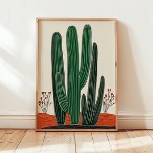 May include: A framed art print featuring a stylized illustration of green cacti with vertical white lines. The cacti are set against a cream background with a red-orange base and small red floral accents. The frame is a light wood color.