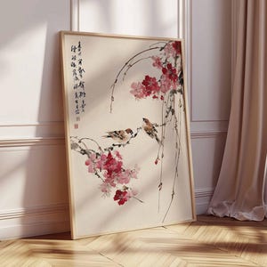 Peut inclure: Une impression d'art encadr&eacute;e repr&eacute;sentant une peinture d'inspiration asiatique. L'&oelig;uvre d'art montre deux oiseaux perch&eacute;s sur une branche fleurie avec des fleurs rouges. De la calligraphie est visible. Le cadre est en bois clair.
