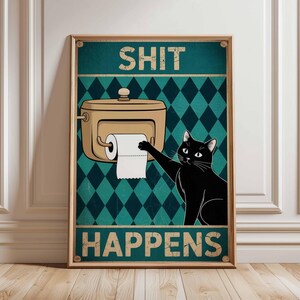 May include: A framed poster with a humorous design. It features a black cat reaching for toilet paper from a dispenser, with the words "SHIT HAPPENS" in bold letters. The background has a teal diamond pattern.