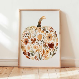 May include: A framed watercolor print featuring a pumpkin shape composed of autumn-colored flowers and foliage. The artwork has a light wooden frame and is displayed against a white background. The color palette includes shades of orange, brown, and green.