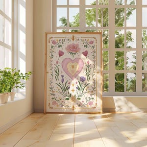 May include: A framed art print featuring a heart design with floral and botanical elements in soft pink, purple, and gold tones. The artwork is displayed in a bright room with a window and plants.