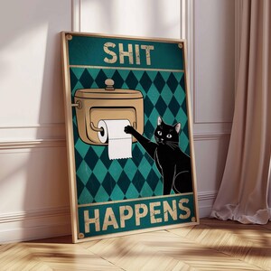 May include: A framed poster with the text "SHIT HAPPENS" and a cartoon image of a black cat reaching for toilet paper. The background is teal with a diamond pattern, and the frame is a light brown color.