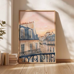 May include: Watercolour painting of Parisian buildings with blue rooftops and balconies, set against a soft pink and orange sunset sky. The artwork is framed in a light wood frame and leans against a wall.