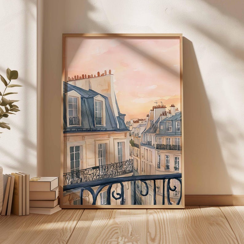 May include: Watercolour painting of Parisian buildings with blue rooftops and balconies, set against a soft pink and orange sunset sky. The artwork is framed in a light wood frame and leans against a wall.