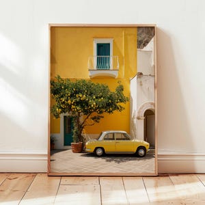 May include: Framed print of a yellow building with a teal door and balcony, a lemon tree, and a yellow vintage car. The building has a white section and a brown wooden frame. The scene is set on a brick patio.