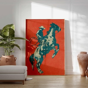 May include: A framed art print featuring a teal and cream-colored cowboy riding a rearing horse against a vibrant red background. The artwork is displayed in a room with a neutral color palette, a potted plant, and a gray ottoman.
