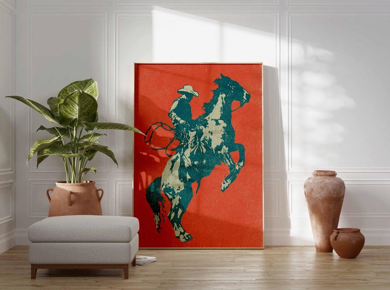 May include: A framed art print featuring a teal and cream-colored cowboy riding a rearing horse against a vibrant red background. The artwork is displayed in a room with a neutral color palette, a potted plant, and a gray ottoman.