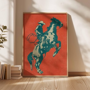 May include: Framed art print featuring a cowboy riding a rearing horse. The artwork uses a teal and beige color palette against a red background. The cowboy is wearing a hat and holding a lasso. The print is in a wooden frame.