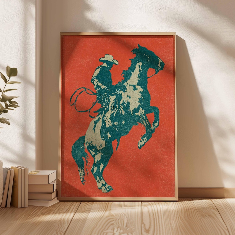 May include: Framed art print featuring a cowboy riding a rearing horse. The artwork uses a teal and beige color palette against a red background. The cowboy is wearing a hat and holding a lasso. The print is in a wooden frame.