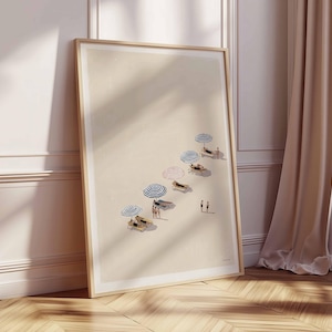 May include: A framed art print showing a beach scene with several beach umbrellas in blue, red, and white. People are relaxing on sun loungers under the umbrellas. The artwork is set against a neutral background, with a wooden frame.