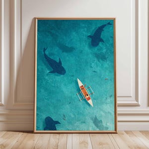 May include: An artistic print depicts a serene ocean scene with a small boat and several dark blue whale sharks swimming beneath the surface. The artwork is framed in a wooden frame and set against a white wall and wooden floor.