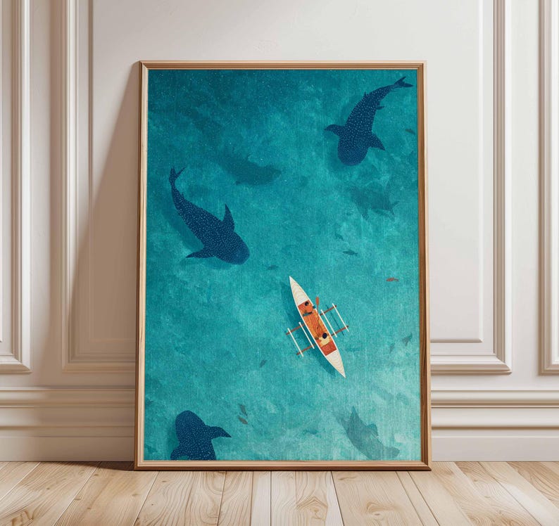 May include: An artistic print depicts a serene ocean scene with a small boat and several dark blue whale sharks swimming beneath the surface. The artwork is framed in a wooden frame and set against a white wall and wooden floor.