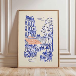 May include: A framed art print depicting a Parisian cafe scene. The illustration features a blue and orange color scheme, with a cafe awning, tables, and buildings. The print is in a wooden frame and is displayed against a cream-colored background.