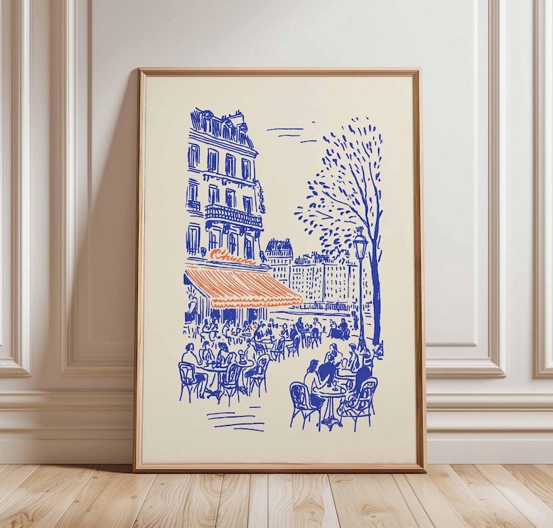 May include: A framed art print depicting a Parisian cafe scene. The illustration features a blue and orange color scheme, with a cafe awning, tables, and buildings. The print is in a wooden frame and is displayed against a cream-colored background.