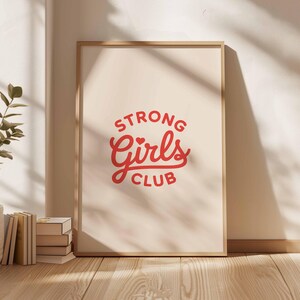 Pu&ograve; includere: Una stampa incorniciata con la scritta "STRONG Girls CLUB" in rosso su sfondo crema. La stampa &egrave; in una cornice di legno chiaro e appoggiata a un muro. Sono visibili libri e una pianta.