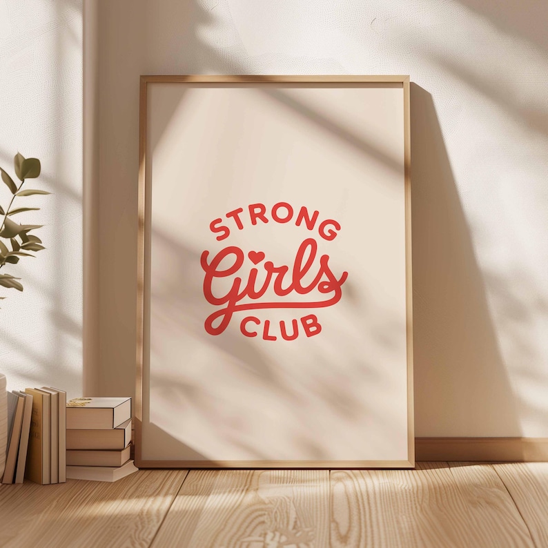 Pu&ograve; includere: Una stampa incorniciata con la scritta "STRONG Girls CLUB" in rosso su sfondo crema. La stampa &egrave; in una cornice di legno chiaro e appoggiata a un muro. Sono visibili libri e una pianta.