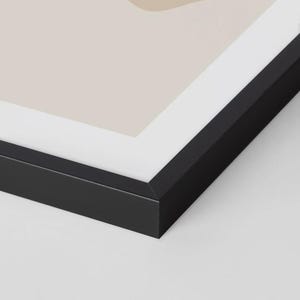May include: A framed abstract art print with a minimalist design. The artwork features beige and tan shapes and black line art on a light background. The frame is black.