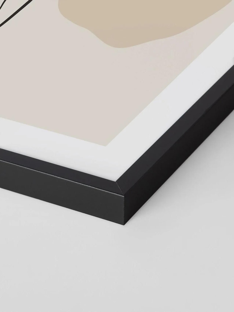 May include: A framed abstract art print with a minimalist design. The artwork features beige and tan shapes and black line art on a light background. The frame is black.
