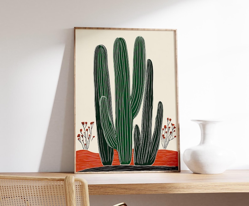 May include: A framed art print featuring a stylized illustration of green cacti against a cream background. The cacti are set on a red-orange ground with small red flowering plants. The frame is a light brown color.