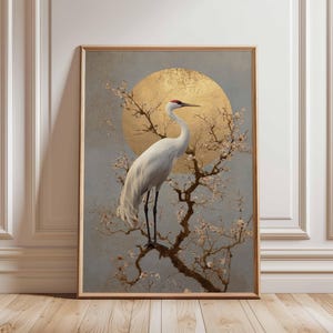 Può includere: Stampa artistica incorniciata raffigurante una gru bianca con una corona rossa posata su un ramo fiorito. Lo sfondo presenta un sole dorato su uno sfondo blu-grigio. La cornice è color legno chiaro.