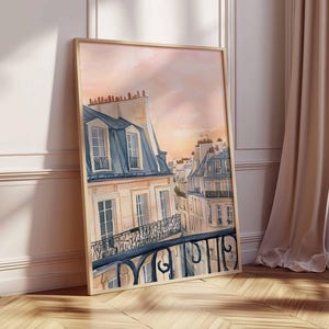 May include: A watercolour painting depicting Parisian rooftops at sunset. The artwork shows buildings with blue-grey mansard roofs, cream walls, and wrought-iron balconies. The sky transitions from peach to pink, creating a warm atmosphere. The framed art leans against a wall.
