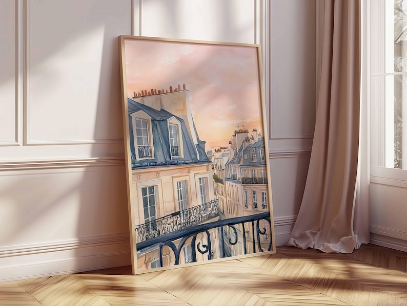 May include: A watercolour painting depicting Parisian rooftops at sunset. The artwork shows buildings with blue-grey mansard roofs, cream walls, and wrought-iron balconies. The sky transitions from peach to pink, creating a warm atmosphere. The framed art leans against a wall.