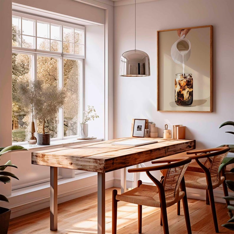 May include: A bright, airy room with a wooden desk and two chairs. A framed picture of iced coffee hangs on the wall. A large window offers a view of trees. A silver pendant light hangs above the desk.