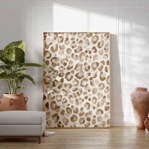 May include: A large framed artwork featuring a leopard print pattern in shades of brown and beige. The artwork is displayed in a room with a neutral color palette, complemented by a potted plant, a gray ottoman, and terracotta vases.