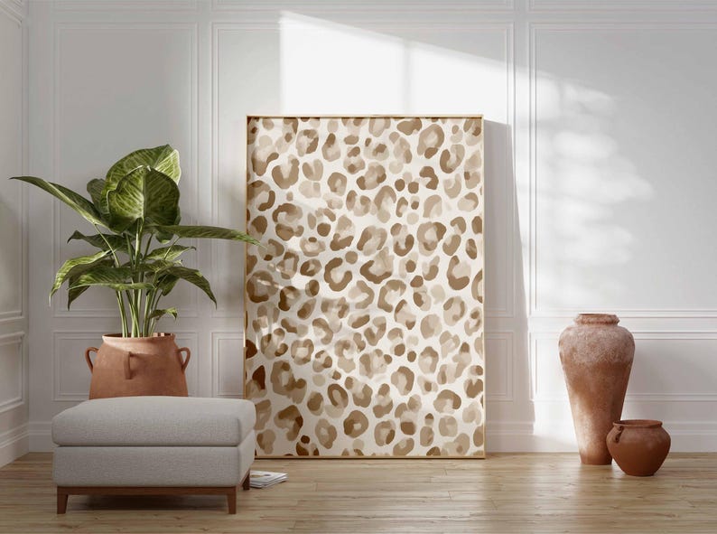 May include: A large framed artwork featuring a leopard print pattern in shades of brown and beige. The artwork is displayed in a room with a neutral color palette, complemented by a potted plant, a gray ottoman, and terracotta vases.