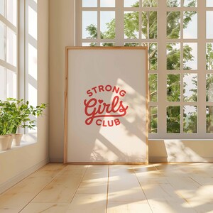 Pu&ograve; includere: Un poster incorniciato con la scritta "STRONG Girls CLUB" in rosso. Il poster &egrave; appoggiato a un muro in una stanza con una grande finestra e pavimenti in legno. Piante in vaso sul davanzale.
