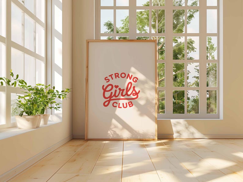 Pu&ograve; includere: Un poster incorniciato con la scritta "STRONG Girls CLUB" in rosso. Il poster &egrave; appoggiato a un muro in una stanza con una grande finestra e pavimenti in legno. Piante in vaso sul davanzale.
