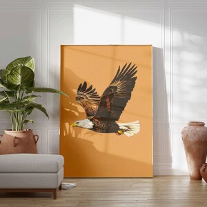 May include: A large framed print featuring a bald eagle in flight against a warm orange background. The eagle has brown and white feathers, a yellow beak, and is positioned to the right. The print is displayed in a room with a plant, ottoman, and decorative vases.