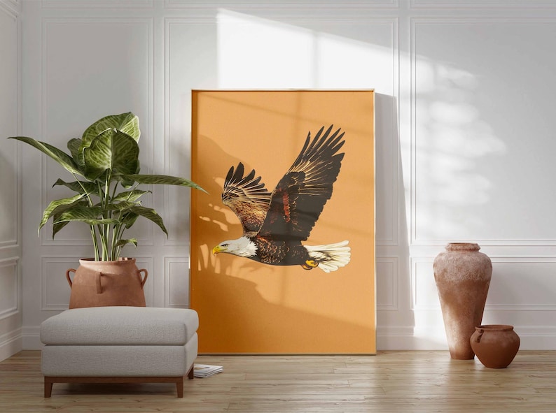 May include: A large framed print featuring a bald eagle in flight against a warm orange background. The eagle has brown and white feathers, a yellow beak, and is positioned to the right. The print is displayed in a room with a plant, ottoman, and decorative vases.