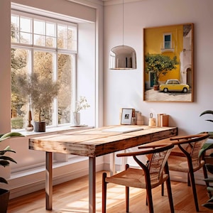 May include: A sunlit interior features a rustic wooden desk with metal legs, two woven chairs, and a large window overlooking a tree-filled landscape. A framed picture of a yellow building with a yellow car hangs on the wall. A chrome pendant light hangs above the desk.