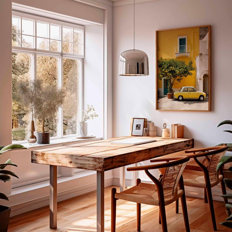 May include: A sunlit interior features a rustic wooden desk with metal legs, two woven chairs, and a large window overlooking a tree-filled landscape. A framed picture of a yellow building with a yellow car hangs on the wall. A chrome pendant light hangs above the desk.