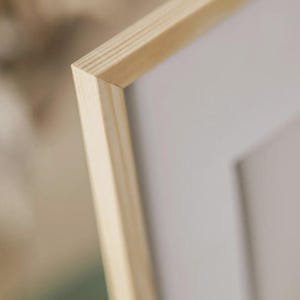 May include: Close-up of a light-coloured wooden picture frame. The frame has a natural wood grain and a clean, modern design. The corner of the frame is visible, showing the precise joinery. The background is blurred, highlighting the frame's details.