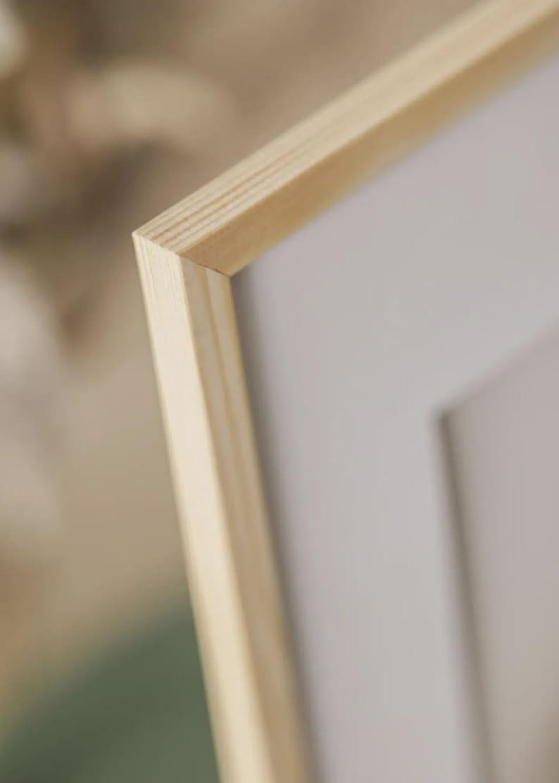 May include: Close-up of a light-coloured wooden picture frame. The frame has a natural wood grain and a clean, modern design. The corner of the frame is visible, showing the precise joinery. The background is blurred, highlighting the frame's details.