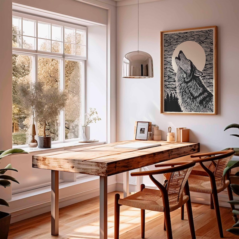May include: A wooden desk with metal legs and two woven chairs in a bright room. A framed black and white print of a howling wolf hangs on the wall. A large window offers a view of trees and natural light.