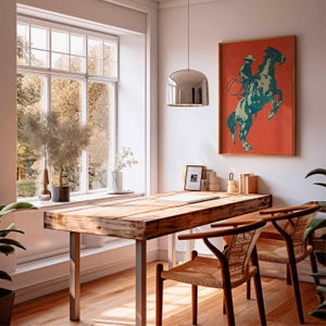 May include: A rustic wooden desk with metal legs and two woven chairs. A framed print of a cowboy on a rearing horse hangs on the wall. The room has a large window and a modern pendant light.
