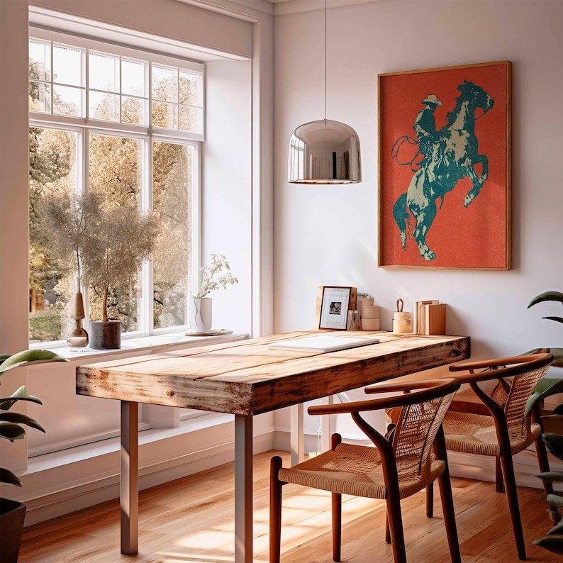 May include: A rustic wooden desk with metal legs and two woven chairs. A framed print of a cowboy on a rearing horse hangs on the wall. The room has a large window and a modern pendant light.