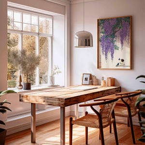 May include: A bright, airy home office with a large wooden desk and two woven chairs. A framed print of a Japanese painting with purple wisteria hangs on the wall. A large window offers a view of the outdoors, and a silver pendant light hangs from the ceiling.