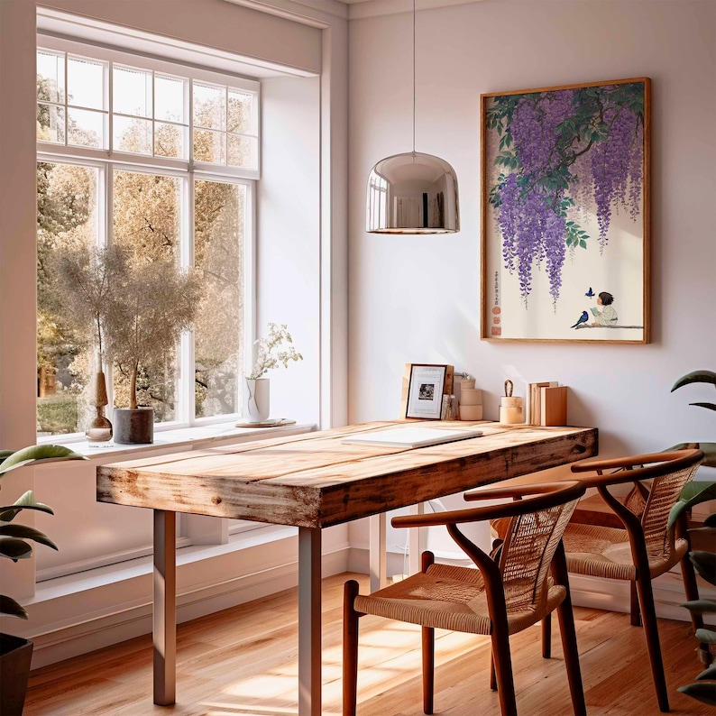 May include: A bright, airy home office with a large wooden desk and two woven chairs. A framed print of a Japanese painting with purple wisteria hangs on the wall. A large window offers a view of the outdoors, and a silver pendant light hangs from the ceiling.