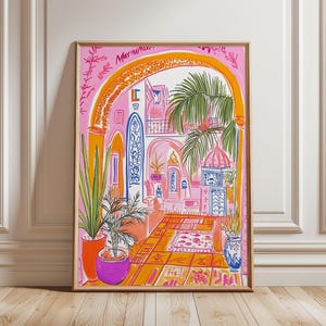 May include: A framed art print depicting a vibrant, colorful courtyard scene. The artwork features pink, orange, and blue hues, with architectural details, potted plants, and a patterned rug. The print is in a wooden frame and leans against a white wall.