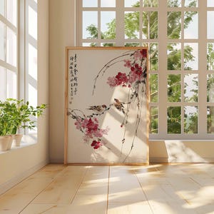 Peut inclure: Une &oelig;uvre d'art encadr&eacute;e repr&eacute;sentant une peinture de style asiatique. Elle montre deux oiseaux perch&eacute;s sur des branches fleuries roses et des baies rouges. Le fond est beige clair, et le cadre est marron clair. La pi&egrave;ce est &eacute;clair&eacute;e par une fen&ecirc;tre.