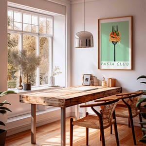 Op de afbeelding: Een lichte, luchtige kamer met een houten tafel en twee stoelen. Een ingelijste "Pasta Club" poster hangt aan de muur. Een groot raam biedt uitzicht op bomen. Een chromen hanglamp hangt boven de tafel.
