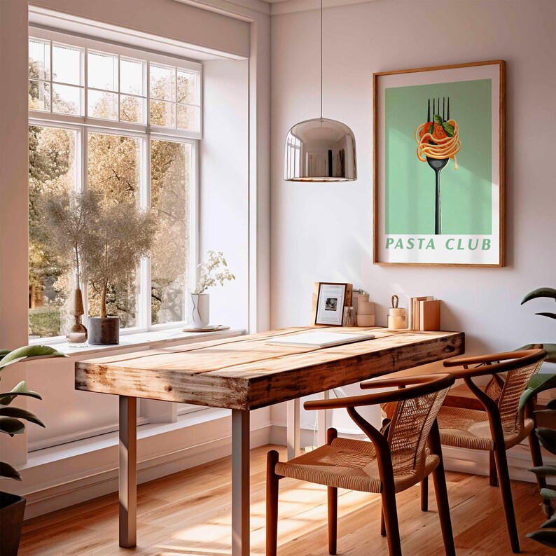 Op de afbeelding: Een lichte, luchtige kamer met een houten tafel en twee stoelen. Een ingelijste "Pasta Club" poster hangt aan de muur. Een groot raam biedt uitzicht op bomen. Een chromen hanglamp hangt boven de tafel.