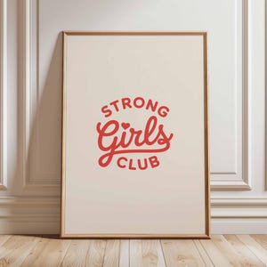 Pu&ograve; includere: Una stampa incorniciata con la scritta "STRONG Girls CLUB" in rosso su sfondo crema. La stampa &egrave; in una cornice di legno e poggia su un muro bianco e un pavimento in legno.