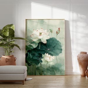 Puede incluir: Una pintura de acuarela enmarcada de flores de loto blancas con grandes hojas verdes. La obra de arte se exhibe en una habitaci&oacute;n con una planta en maceta, un otom&aacute;n gris y jarrones de terracota decorativos. La habitaci&oacute;n tiene paneles blancos y un suelo de madera.