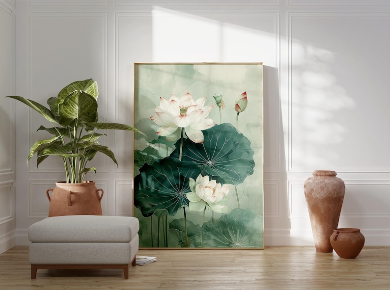Puede incluir: Una pintura de acuarela enmarcada de flores de loto blancas con grandes hojas verdes. La obra de arte se exhibe en una habitaci&oacute;n con una planta en maceta, un otom&aacute;n gris y jarrones de terracota decorativos. La habitaci&oacute;n tiene paneles blancos y un suelo de madera.